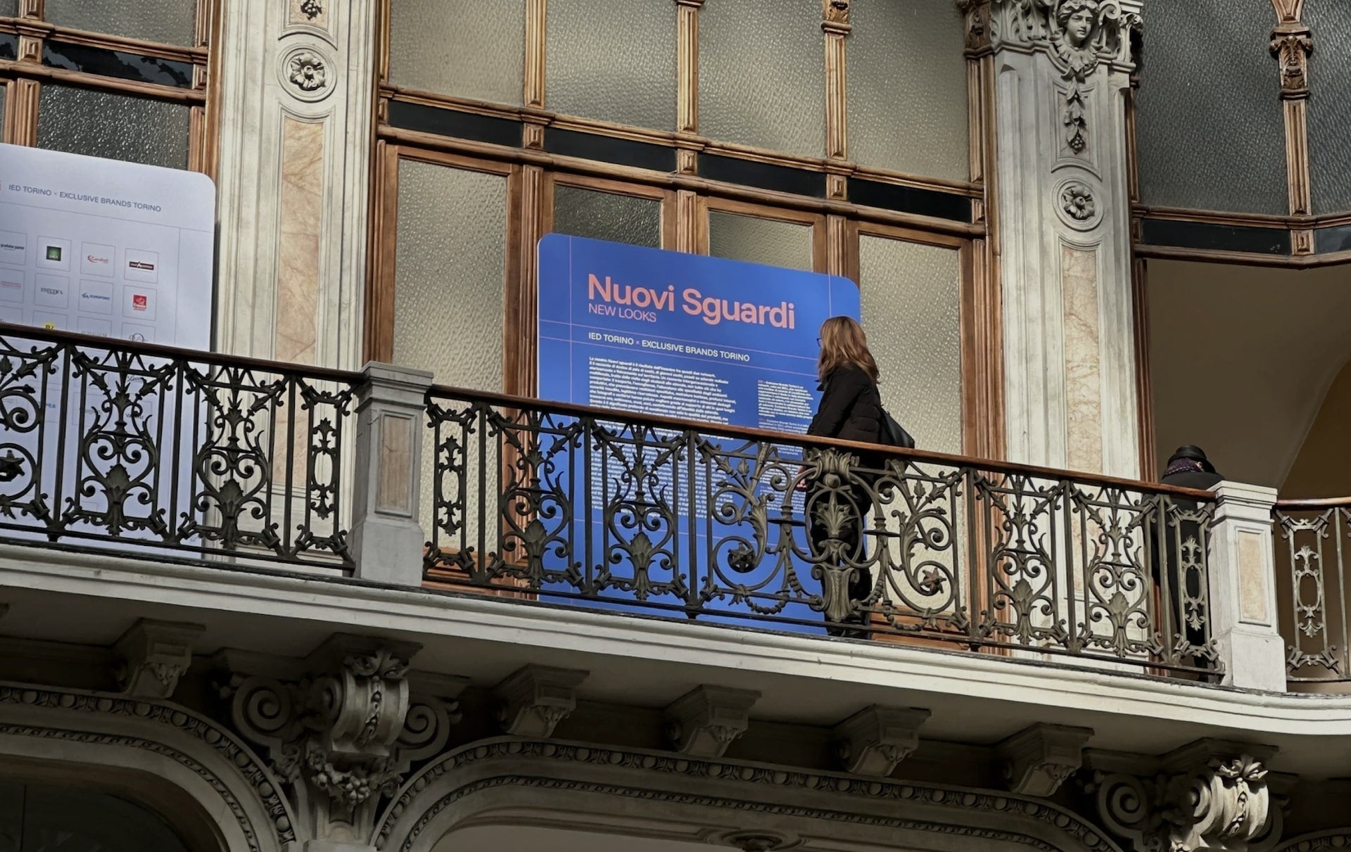 Person standing on a balcony with wrought iron railing, observing the exhibition’s informational panel
