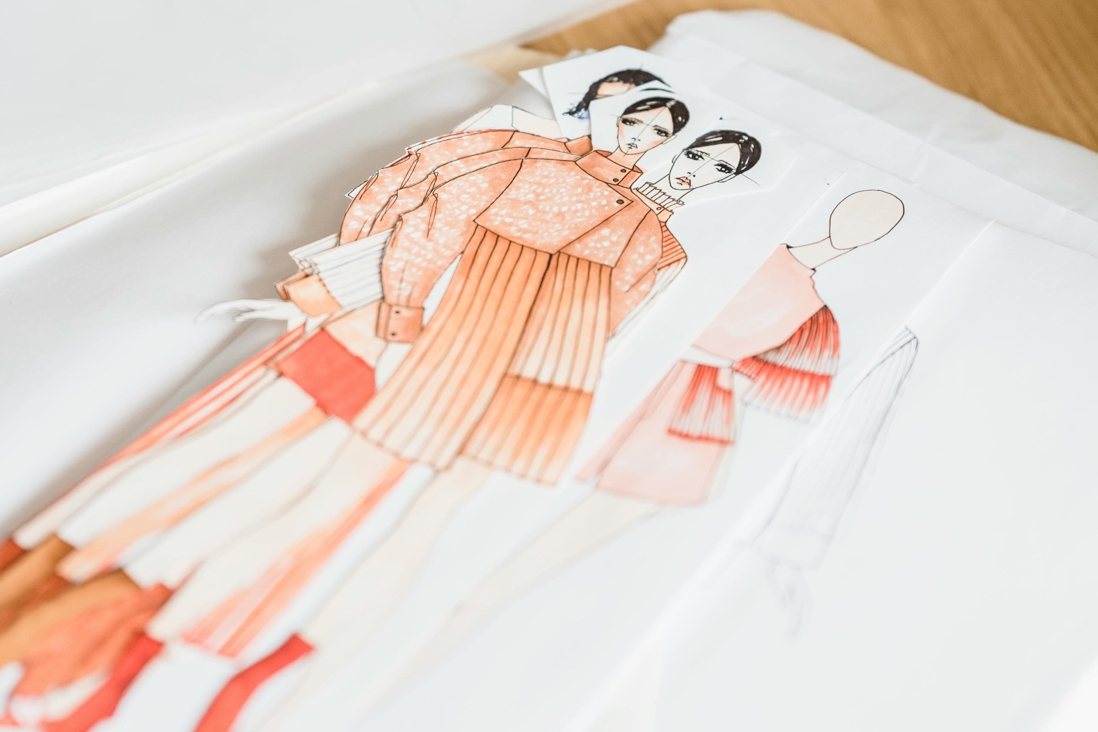 Fashion sketches on paper featuring female figures in orange outfits with pleated details, laid out on a work table