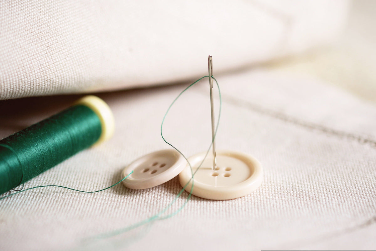 Sewing needle, green thread, and buttons resting on a light-colored fabric